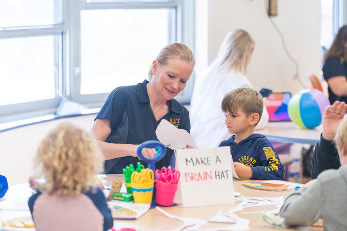 Rain Bosworth interacting with a child sitting next to her at the National Technical Institute for the Deaf. Bosworth's blonde hair is pulled back into a ponytail, and she's wearing a short-sleeve navy shirt that displays the RIT logo in orange. The child. who has short blonde hair and wearing a navy hoodie, is watching Bosworth cutting a piece of paper with a pair of scissors. They are both sitting at a round table, which has a sign that reads, "Make a brain hat."