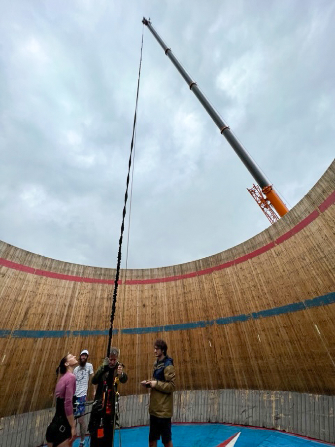 Four people stand in the middle of a cylindrical structure, looking up at a cloudy sky, in preparation for a moon running experiment. A long black bungee cord stretches down into the structure from a nearly 40 meter tall crane.