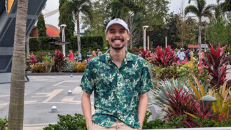 Computer Scientist Niall Williams stands in front of metal handrail. He's has a black moustache and beard. He's wearing a powder blue baseball cap and a button down shirt with tree designs on it. A concrete courtyard and palm trees are in the background.
