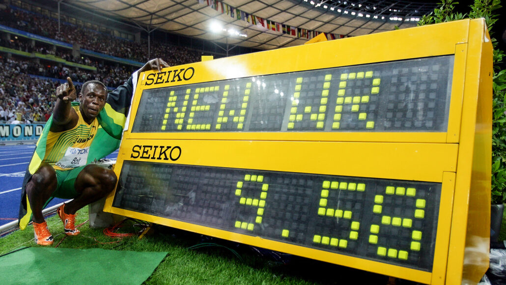 Usain Bolt crouches next to a digital display showing his new world record while pointing to a crowd in a stadium