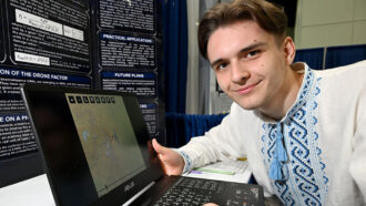 Volodymyr Borysenko is leaning over a laptop showing his research at Regeneron ISEF. He has light brown hair and is wearing a white tunic with blue embroidery around his collar and down the middle of his shirt.