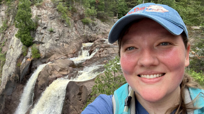 Anne Kort takes a selfie in front of High Falls at Tettegouche State Park in northern Minnesota. She's wearing a blue baseball cap and a teal backpack.