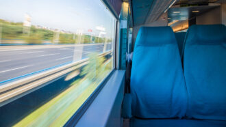 Two plush blue seats are against the window of a dimly lit train car, while outside the view of a highway and blue sky whizzes by so fast it's blurry