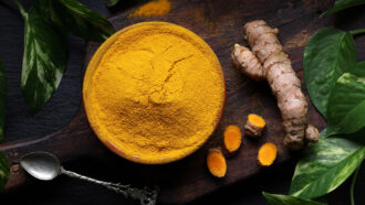 a bowl of bright yellow ground tumeric on a table top next to a tumeric root