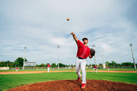 A baseball pitcher wearing a red jersey and white pants pitches a ball from a brown mound. A green baseball field surrounds him below cloudy skies