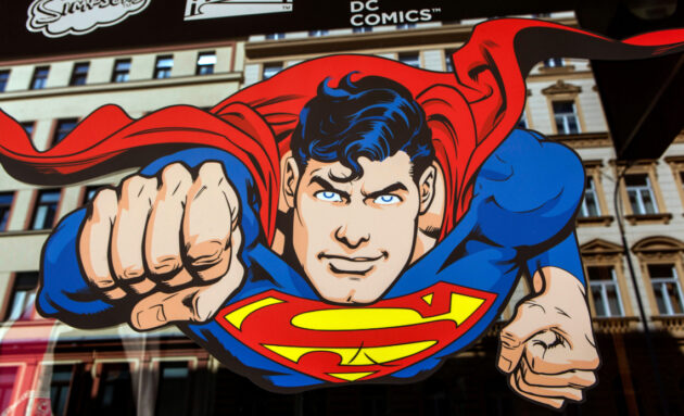 An illustrated Superman flies head on toward the viewer. He is a white man with black hair and blue eyes. His costume sports a yellow diamond with a red S in the middle. His red cape trails behind him. A tan, multistory building is in the background