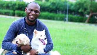 Sunday Agbonika sits in a field with two dogs in his lap. His own tan and white pooch, Tinkerbell is on the left, and his friend’s white dog, Snow is on the right. Agbonika is a Black man with short hair. He wears a long sleeved blue shirt.