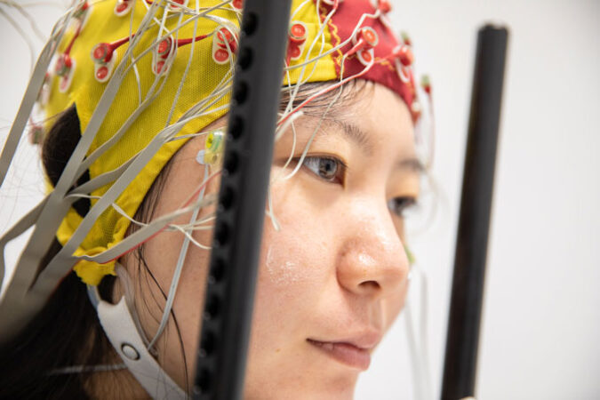 A brain study participant is facing to the right, wearing a yellow and red electroencephalogram (EEG) head gear that's tight around the participant's head like a swim cap. The head gear has electrodes attached to it, and it has a chip strap that goes around the bottom of the participant's face.