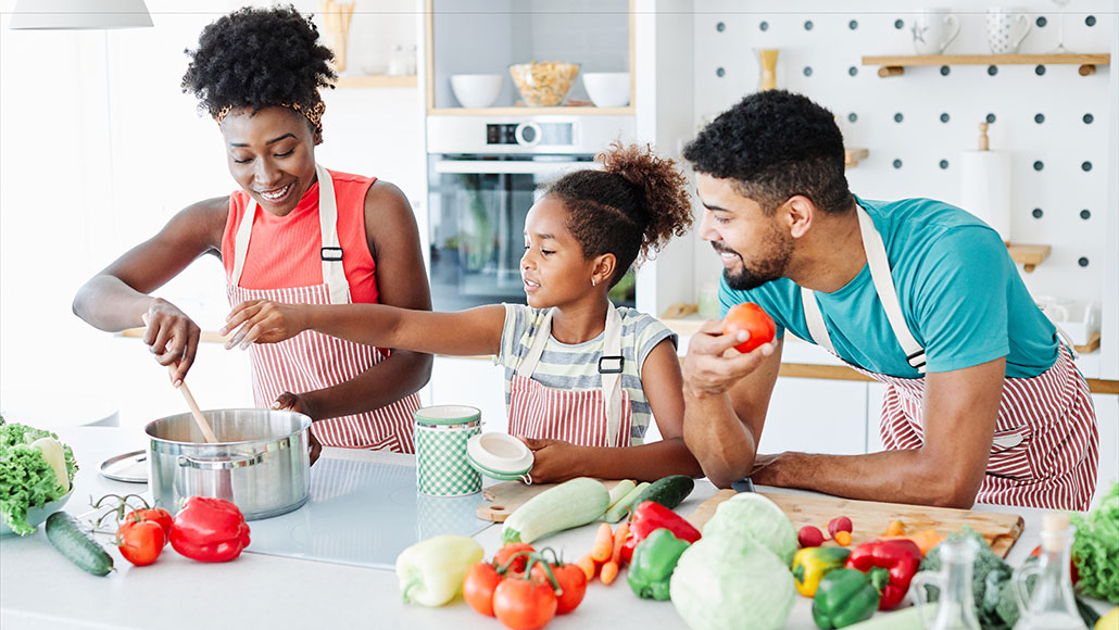 A woman with dark brown skin and curly hair is in the kitchen stirring a pot on the stove while her daughter and husband assist in cooking. The counter is covered in vegetables.