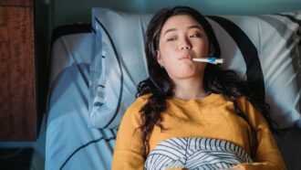 a young Asian woman with long black hair is propped up in bed with a thermometer in her mouth