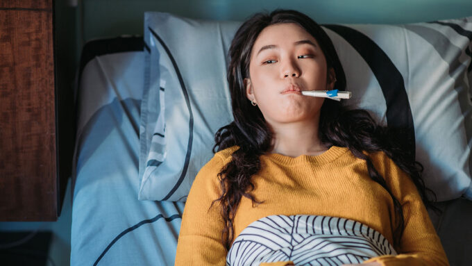 a young Asian woman with long black hair is propped up in bed with a thermometer in her mouth