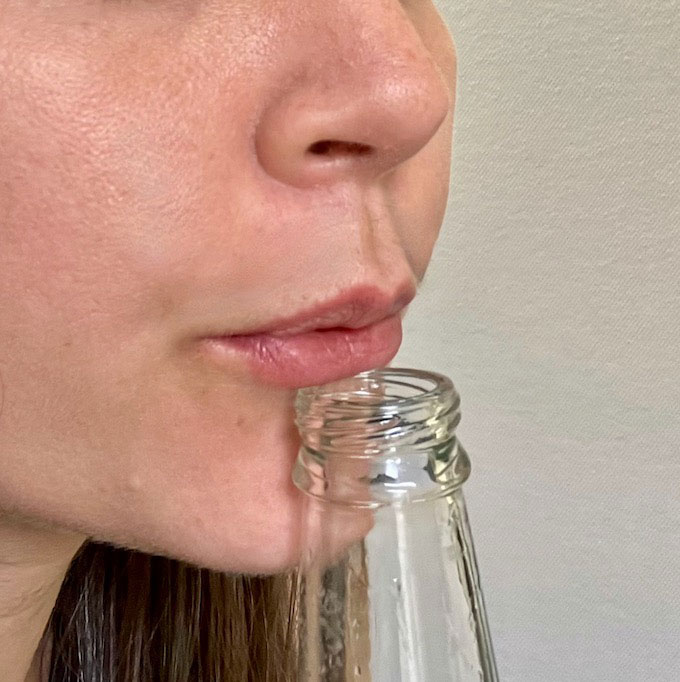 a close-up of a person pursing their lipe to blow across the top of a glass bottle