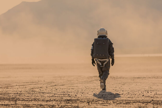 Full length photo of astronaut in space suit and helmet on Mars walking away from the camera towards a dust storm in the distance, red orange all around