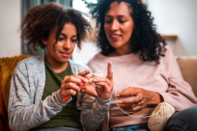 Smiling girl with an afro sits next to an adult teaching her how to crochet