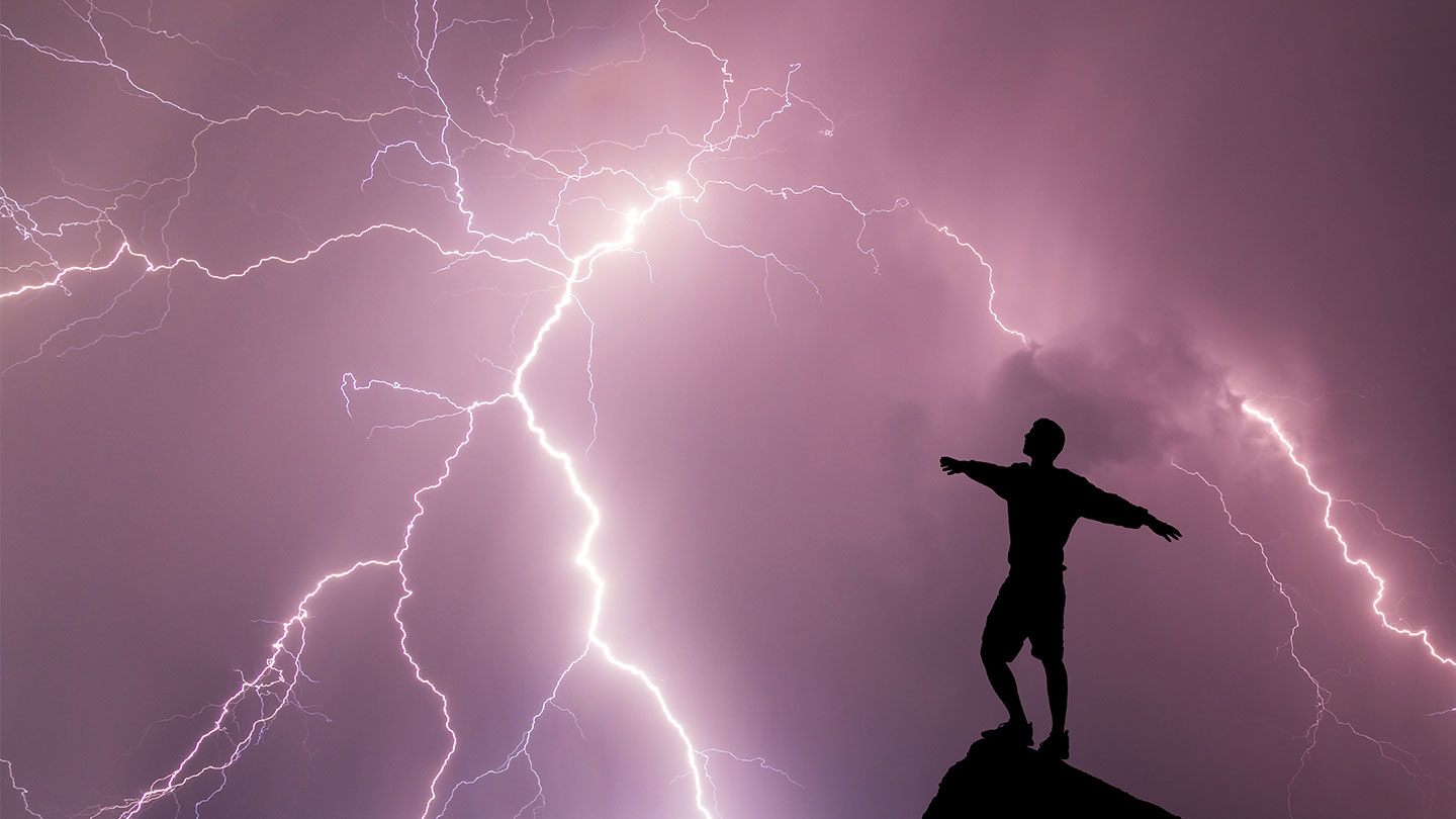 a silhoutte of a person standing on a rocky perch against a purple clouded sky and a lightning strike