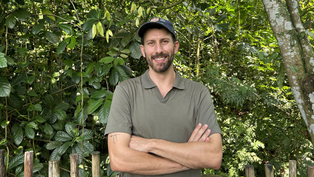 Elias Garcia Pelegrin stands in front of a tropical forest. He has light brown hair and a beard. He is wearing a green polo shirt and matching baseball cap.