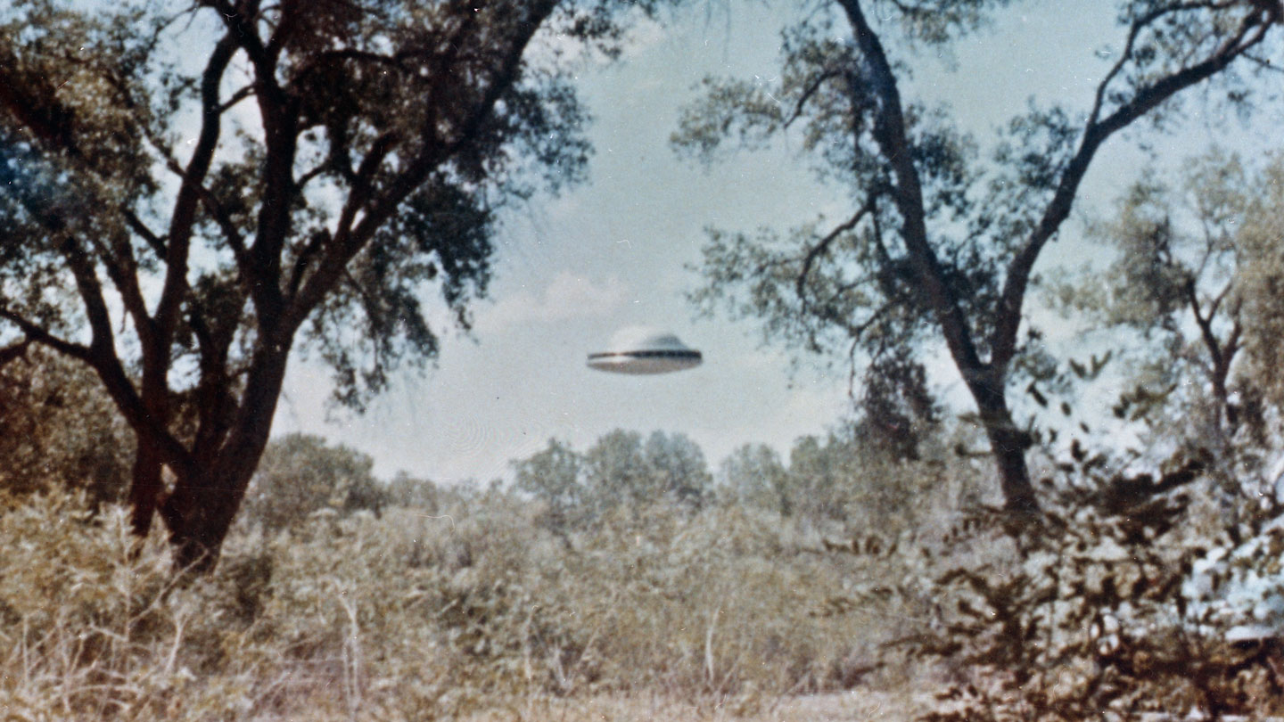 an washed out archival photo showing a silver disc with a half bubble on top floating above a brown field. Trees are on either side of the image.