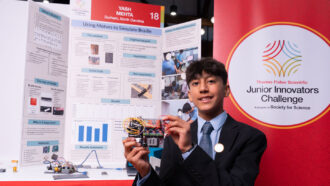 Yash Mehta stands in front of a scientific poster with words and pictures describing his project. The braille reader he holds is an electronic device with several wires and components.