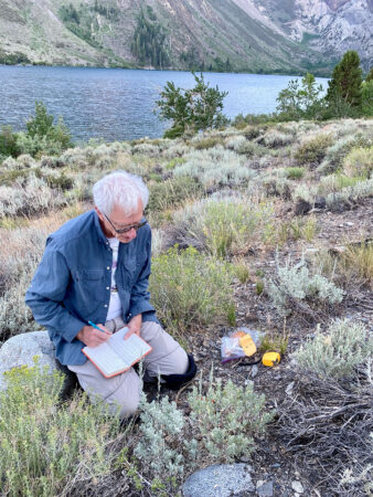 A man in a blue shirt kneels among a scrubby, rocky landscape, a pencil in hand and notebook in his lap. Behind is a lake and a hill.