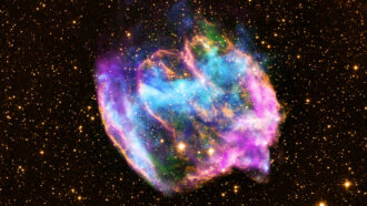 A false-color image of a supernova remnant includes clouds of blue, green, pink and yellow against a backdrop of stars