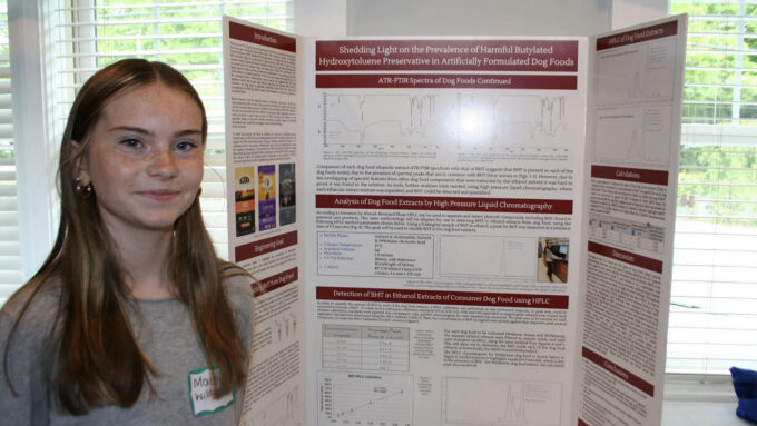 a photo of Mackensey Wilson, a young girl with pale skin, freckles and straight hair standing next to her science project poster board about finding BHT in pet food