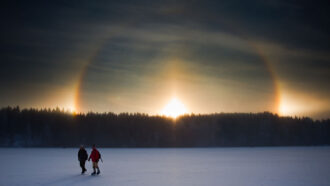 Two people walk on a frozen surface. The patterned glow of a sundog appears on the horizon.