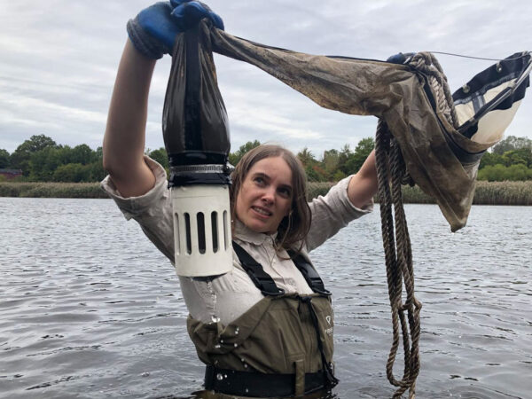 Sally Warring holds a plankton net above her head. She's wearing green waders, standing in a reservoir. Green foliage can be seen behind her. 