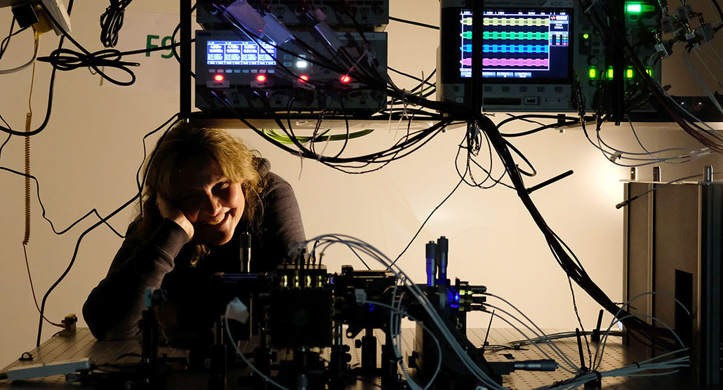 a photo of s smiling lady with her head propped up on one hand, next to an array of cords and computer equipment