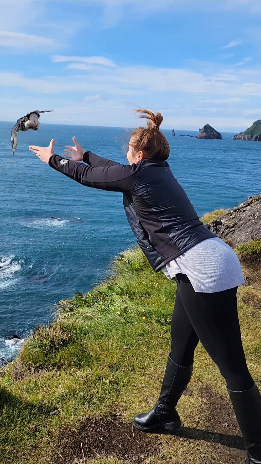 a woman tosses a puffling up into the air on a cliffside above the water