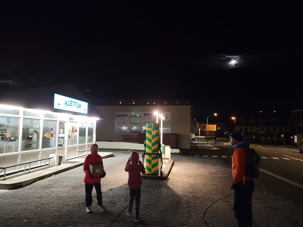 a photo of a gas station with bright lights at night, and the moon in the distance above as seen through clouds