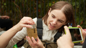 Biologist Sally Warring examines a jar containing pond water. She is a white woman with brown, straight hair and fair skin.