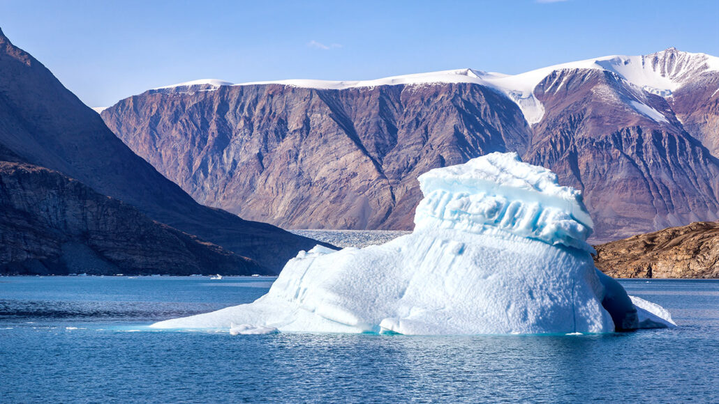 a photo of Dickson Fjord, a small iceberg is in ocean water close to steep snow covered cliffs