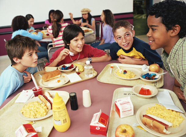 School aged kids sit around a cafeteria table holding lunch trays of food.