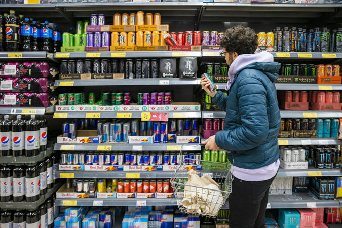 A man wearing a blue puffer jacket reads a food label in front of shelves stocked with food.