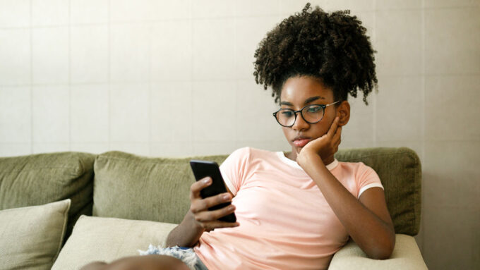 A bored-looking woman sits on a tan couch scrolling through her smartphone.