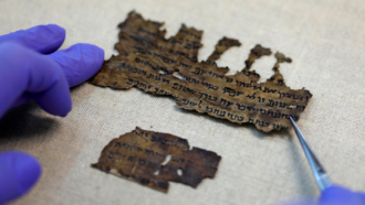 gloved hands and forceps touch darkened scraps of ancient scrolls with visible writing, which have been studied with carbon dating