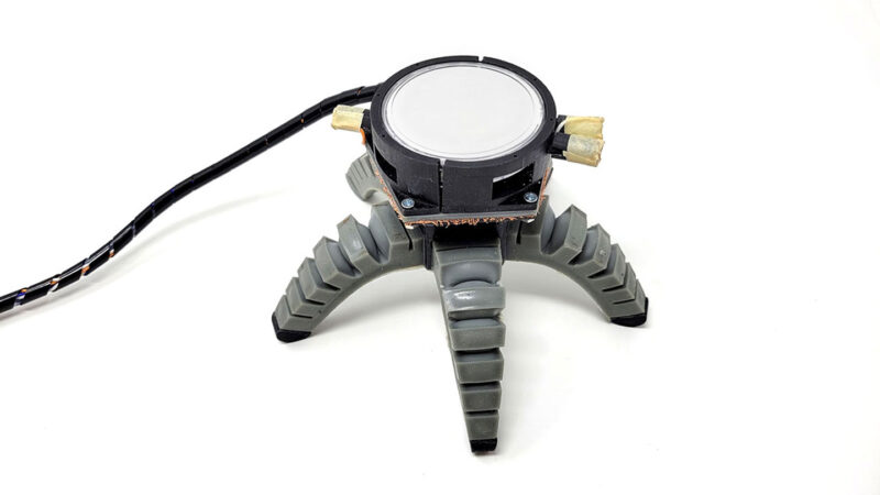 A biohybrid bot with a circular light sensor stands on a white surface. It has five grey legs and a black power cable running to its body.