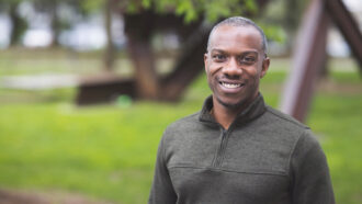 Physicist James Whitfield smiles at the camera. Green grass and a single tree can be seen in the background.He is a Black man with brown eyes. He's wearing a pine green sweater.