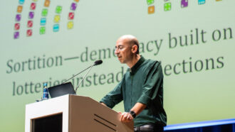 Computer scientist Ariel Procaccia stands at a podium. A projected image with words and colorful blocks is visible behind him.