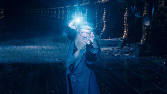 Fictional wizard Albus Dumbledore holds up a wooden wand, perhaps to cast a memory spell. A bright bluish-white light shining from the tip casts a blue glow over the chamber behind him.