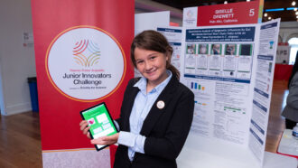 A smiling 12-year-old girl with brown hair and wearing a suit poses with an iPad and poster showcasing her research about dyslexia and diet