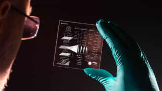 a photo of a white man wearing glasses as seen from behind, he is backlit and holding a large square piece of glass with diagrams and other data