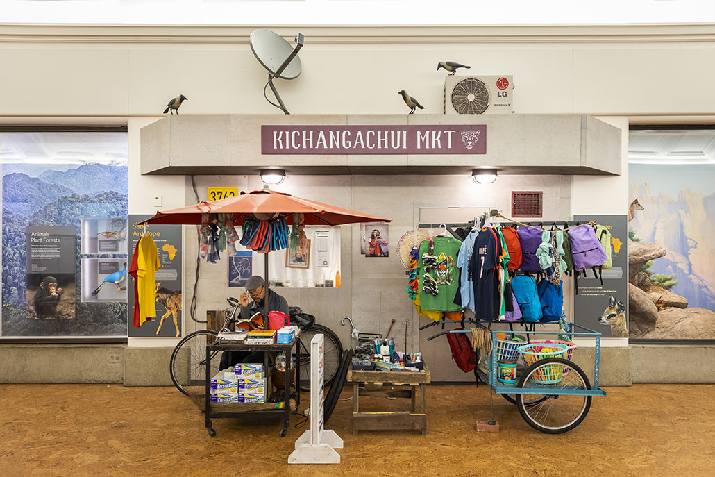 a photo of a display showing a small market storefront, an umbrella covers the storeowner (a person dressed the part) and various goods are displayed on racks, shelves and even a turquiose blue bike wagon. The sign above the stall reads "KICHANGACHUI MKT"