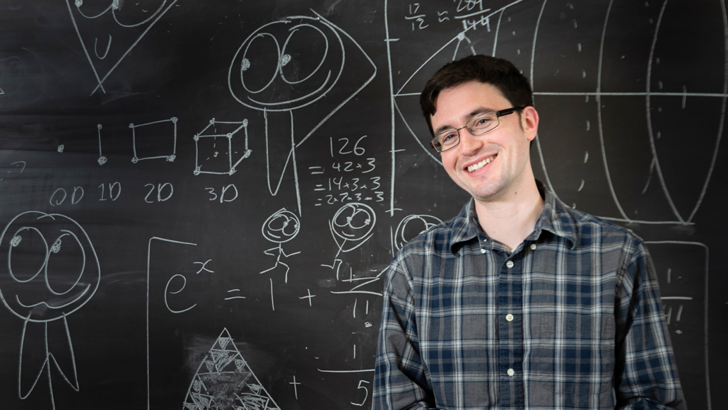 A white man with dark hair and glasses is smiling and standing in front of a chalkboard full of cute drawings and math formulas