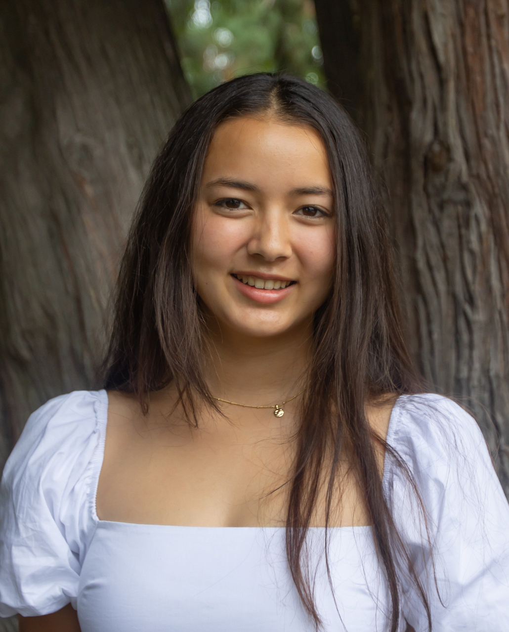 a teen girl with brown hair and brown eyes, wearing a bluish white dress stands beneath a tree